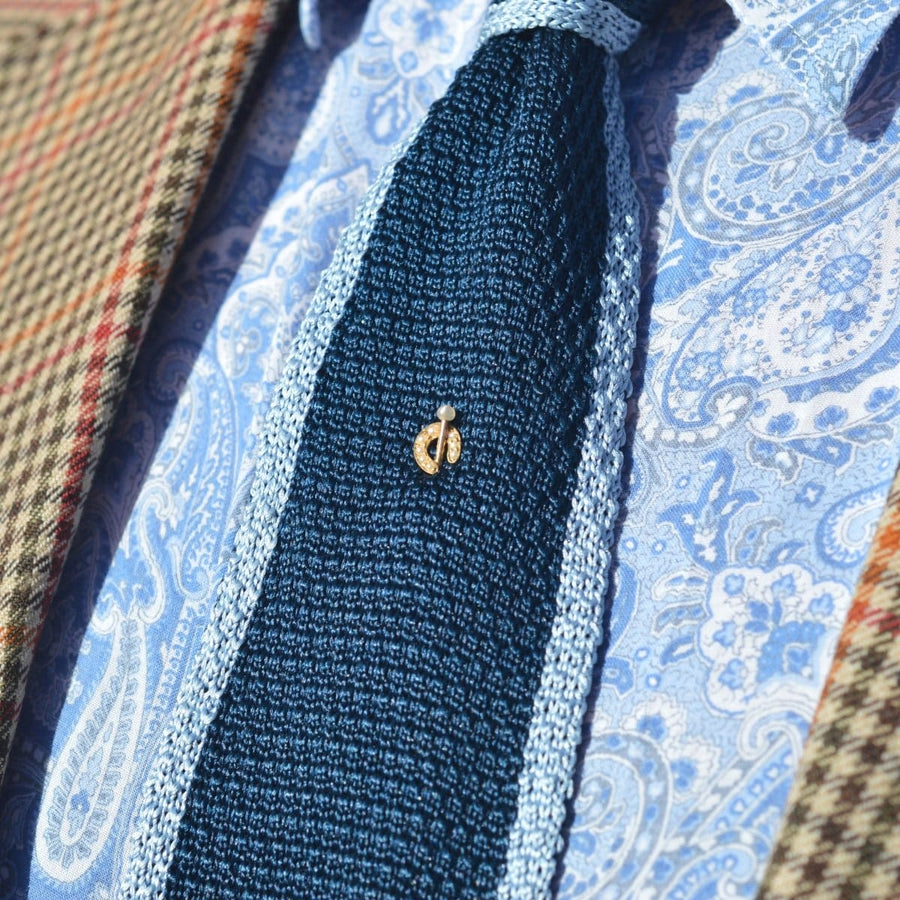 Close-up of a stylish blue tie with a gold horseshoe pin against a patterned shirt and blazer.
